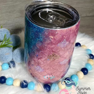 May include: A stemless wine tumbler with a mermaid scale design. The tumbler features a gradient of blue and pink glitter, with a clear lid. The design includes glitter accents and a floral embellishment. The tumbler is on a white fluffy surface.