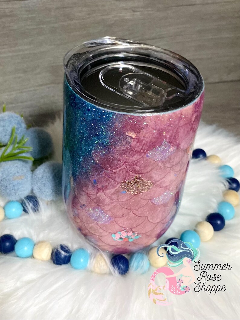 May include: A stemless wine tumbler with a mermaid scale design. The tumbler features a gradient of blue and pink glitter, with a clear lid. The design includes glitter accents and a floral embellishment. The tumbler is on a white fluffy surface.