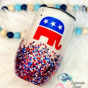 May include: A white glitter wine tumbler featuring a red, white, and blue elephant logo. The lower half of the tumbler is decorated with red, white, and blue glitter. The tumbler has a stemless design.