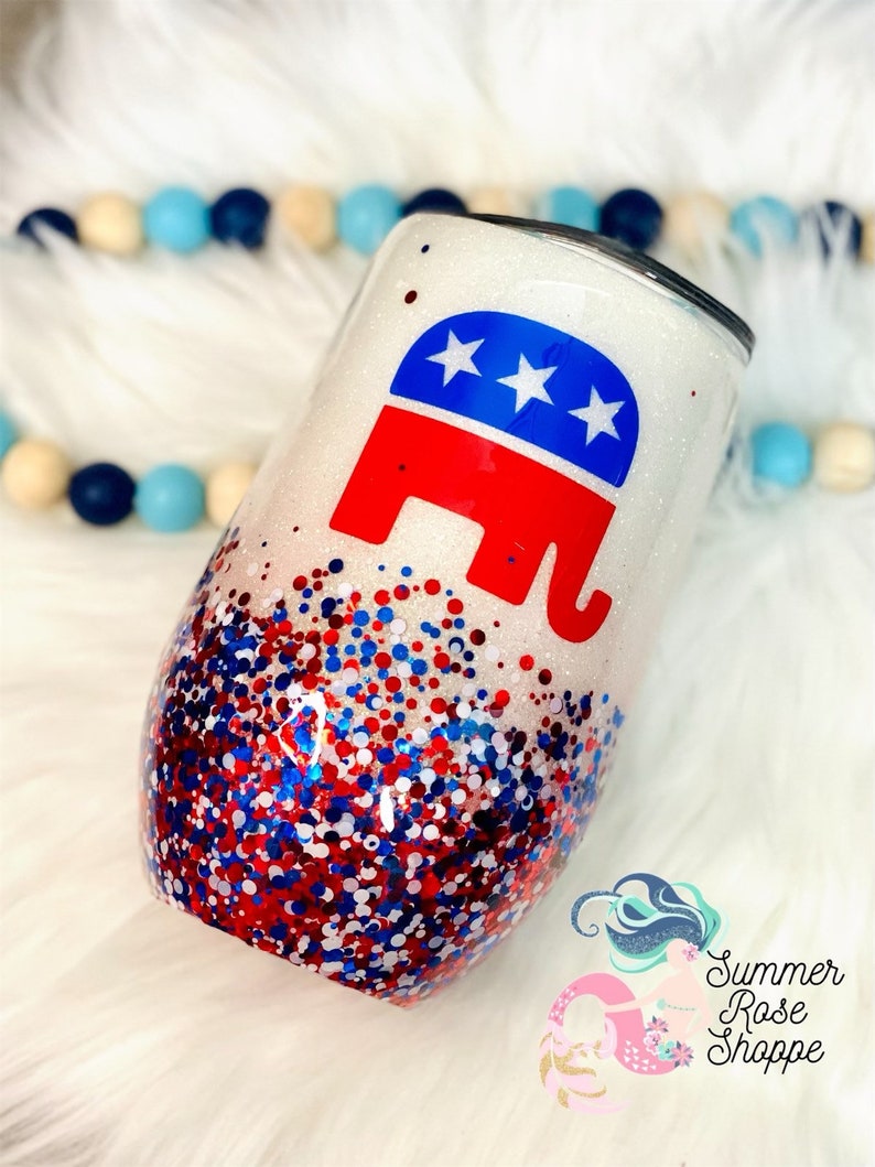 May include: A white glitter wine tumbler featuring a red, white, and blue elephant logo. The lower half of the tumbler is decorated with red, white, and blue glitter. The tumbler has a stemless design.