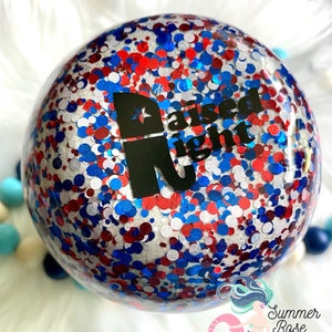 May include: A clear, spherical ornament filled with red, white, and blue glitter. The ornament features the words "Raised Right" in black. It is set against a backdrop of white fluff and colourful beads.