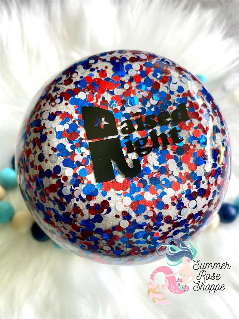 May include: A clear, spherical ornament filled with red, white, and blue glitter. The ornament features the words "Raised Right" in black. It is set against a backdrop of white fluff and colourful beads.