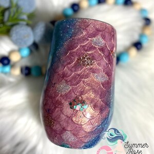 May include: A stemless wine tumbler with a mermaid scale design. The tumbler features a gradient of colors, including shades of purple, blue, and glitter accents. The design is reminiscent of a mermaid's tail. The tumbler is a unique and decorative item.
