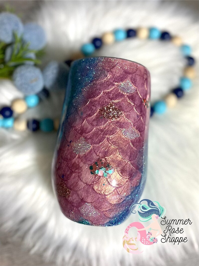 May include: A stemless wine tumbler with a mermaid scale design. The tumbler features a gradient of colors, including shades of purple, blue, and glitter accents. The design is reminiscent of a mermaid's tail. The tumbler is a unique and decorative item.
