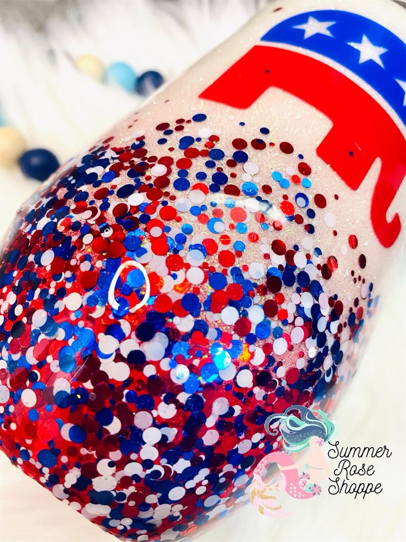 May include: A glitter-covered tumbler with red, white, and blue confetti. The design includes a red elephant graphic with a blue star-studded top. The tumbler is a patriotic theme.