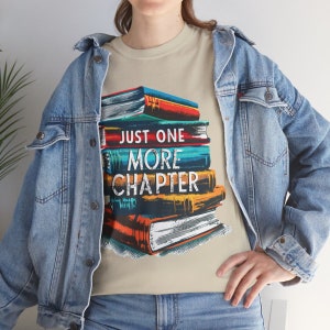 Puede incluir: Una camiseta beige con una pila de libros coloridos y el texto "Just One More Chapter".