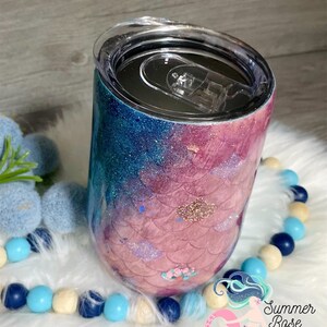 May include: A glitter-covered tumbler with a mermaid scale design. The tumbler transitions from blue to pink, with gold accents. The clear lid has a sliding closure. The tumbler is decorated with a mermaid scale pattern and the text "Summer Rose Shoppe".