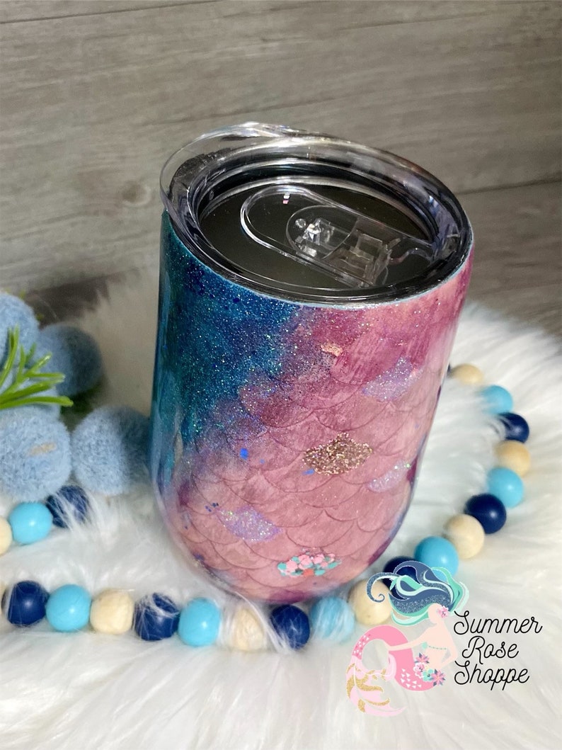 May include: A glitter-covered tumbler with a mermaid scale design. The tumbler transitions from blue to pink, with gold accents. The clear lid has a sliding closure. The tumbler is decorated with a mermaid scale pattern and the text "Summer Rose Shoppe".