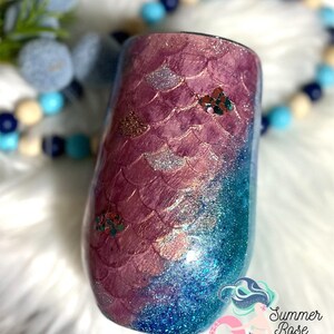 May include: A stemless wine tumbler with a mermaid scale design. The top half features a pink scale pattern, transitioning to a blue glitter base. Embellished with glitter accents and a logo that reads "Summer Rose Shoppe."