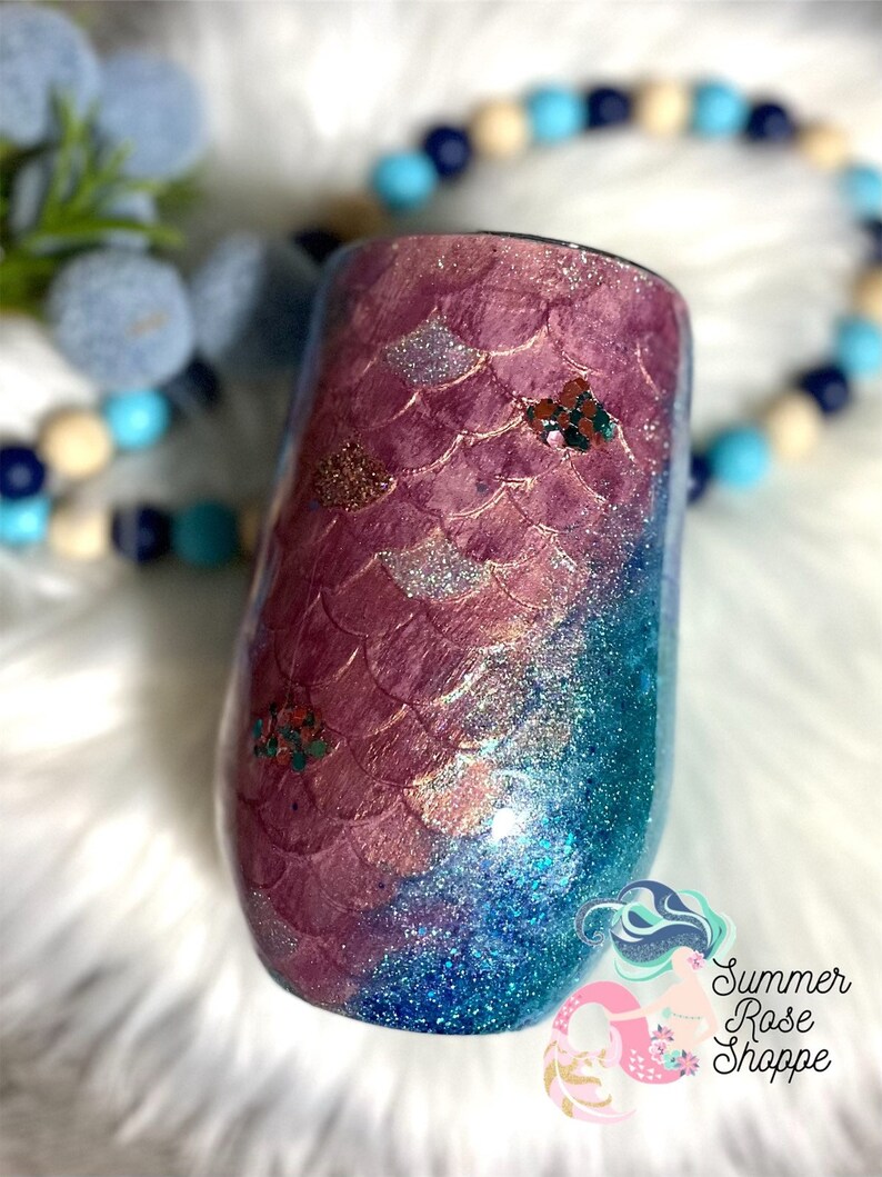May include: A stemless wine tumbler with a mermaid scale design. The top half features a pink scale pattern, transitioning to a blue glitter base. Embellished with glitter accents and a logo that reads "Summer Rose Shoppe."