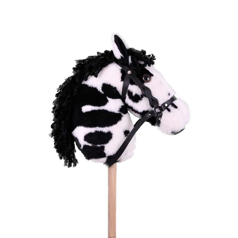May include: A black and white zebra-patterned plush horse head on a wooden stick. The horse has a black leather bridle with a buckle.