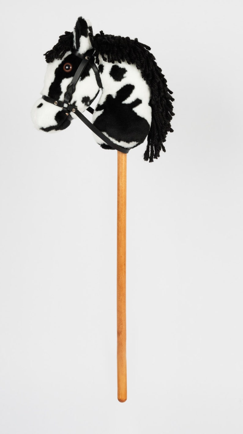 May include: A black and white spotted plush horse head on a wooden stick. The horse has a black mane and a black leather bridle.