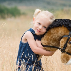 May include: A young girl with blonde hair and blue eyes is hugging a brown and black stuffed horse. She is wearing a blue denim overall dress. The background is a field of tall grass.