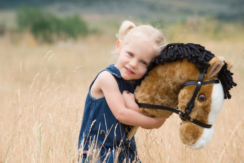 May include: A young girl with blonde hair and blue eyes is hugging a brown and black stuffed horse. She is wearing a blue denim overall dress. The background is a field of tall grass.