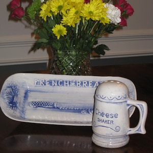 May include: A white ceramic rectangular tray with blue accents featuring the text FRENCH BREAD and a bread knife illustration, paired with a matching white ceramic cheese shaker with blue text and a handle, both decorated with blue trim.