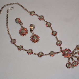 Op de afbeelding: Een vintage sieradenset met een ketting en bijpassende oorbellen. De ketting en oorbellen hebben een bloemmotief met imitatieparels en iriserende kristallen. Het metaal heeft een goudkleurige tint. De oorbellen zijn clip-on.