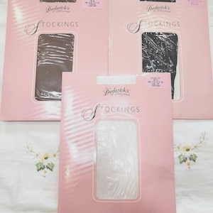 May include: Three packages of Frederick's of Hollywood stockings in brown, black, and white. Each pink rectangular package features the brand name, the word STOCKINGS, and a clear window displaying the hosiery inside. The labels indicate one size fits all.
