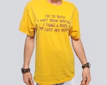 Könnte beinhalten: Senfgelbes T-Shirt mit dem roten Text "I'M SO BUSY I DON'T KNOW WHETHER I FOUND A ROPE OR LOST MY HORSE". Das Shirt wird mit hellblauen zerrissenen Jeans kombiniert. Eine Uhr ist am Handgelenk zu sehen.