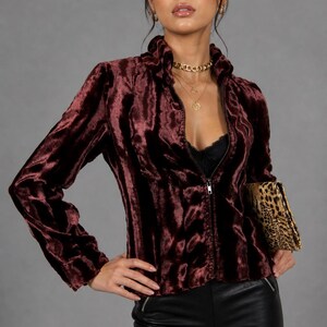 May include: A burgundy velvet zip-up jacket with a high collar. The jacket is paired with black shorts and a leopard print clutch. The model is wearing gold necklaces and earrings. The background is a solid gray.
