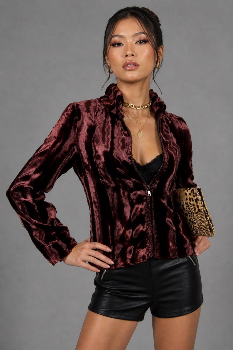 May include: A burgundy velvet zip-up jacket with a high collar. The jacket is paired with black shorts and a leopard print clutch. The model is wearing gold necklaces and earrings. The background is a solid gray.