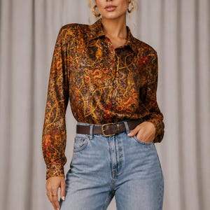 May include: A long-sleeved button-down shirt featuring a complex, multi-colored pattern of gold chains, red floral motifs, and brown swirls. The shirt is tucked into light blue denim jeans secured with a dark brown leather belt with a gold buckle.