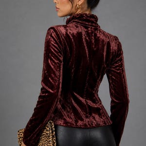 May include: Burgundy velvet jacket with a high collar and long sleeves. The jacket is fitted and has a tailored silhouette. The model is also wearing black shorts and holding a leopard print clutch. The background is a solid gray.