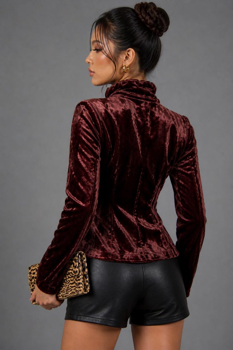 May include: Burgundy velvet jacket with a high collar and long sleeves. The jacket is fitted and has a tailored silhouette. The model is also wearing black shorts and holding a leopard print clutch. The background is a solid gray.