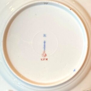 May include: The underside of a white porcelain plate with a blue and red mark that reads "KPM".