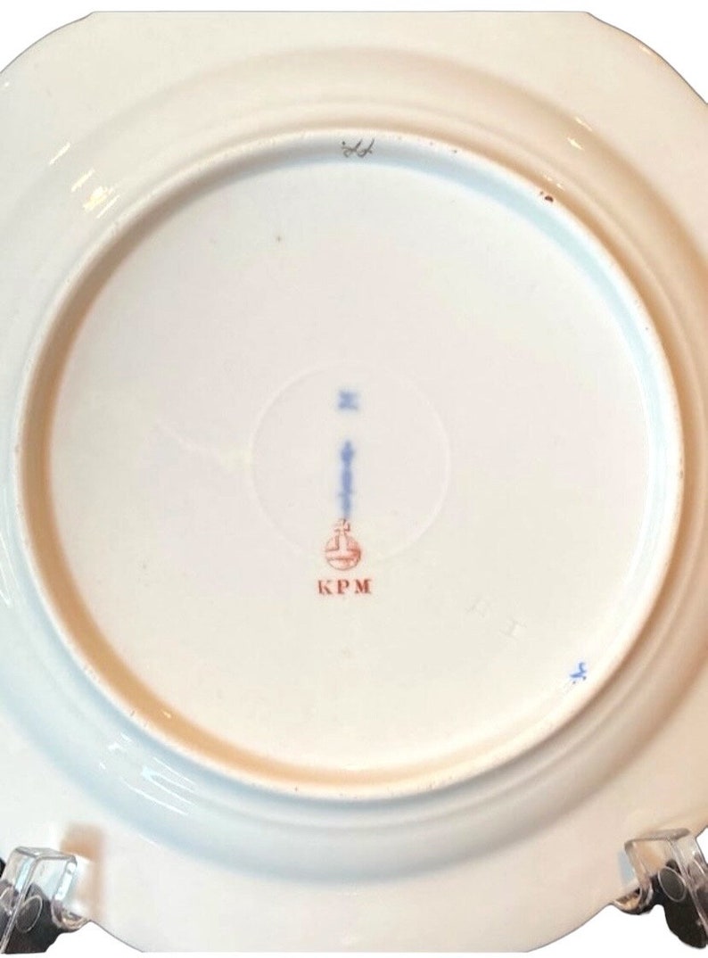 May include: The underside of a white porcelain plate with a blue and red mark that reads "KPM".