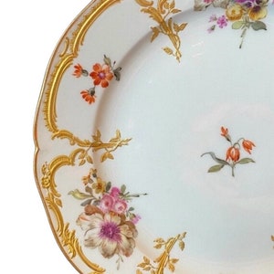 May include: A white porcelain plate with a gold rim and floral pattern. The plate features a variety of flowers in shades of pink, orange, and yellow, with green leaves and stems. The plate has a scalloped edge and a delicate gold design.