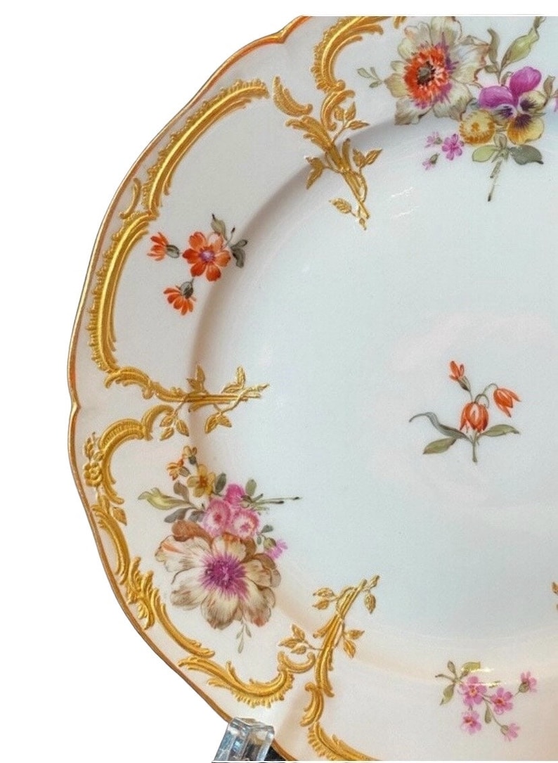 May include: A white porcelain plate with a gold rim and floral pattern. The plate features a variety of flowers in shades of pink, orange, and yellow, with green leaves and stems. The plate has a scalloped edge and a delicate gold design.