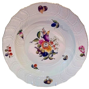 May include: A white porcelain plate with a raised woven pattern around the rim. The center of the plate features a hand-painted floral design with a variety of fruits and vegetables, including an eggplant, apple, and berries.