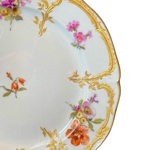 May include: A white porcelain plate with a gold rim and floral pattern. The plate features delicate pink, orange, and yellow flowers with green leaves. The gold rim is intricate and detailed.