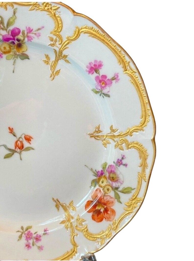 May include: A white porcelain plate with a gold rim and floral pattern. The plate features delicate pink, orange, and yellow flowers with green leaves. The gold rim is intricate and detailed.