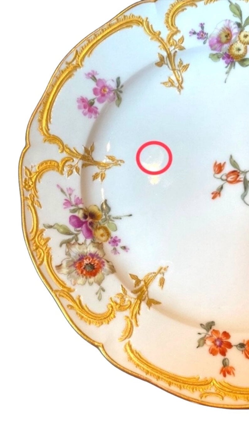 May include: A white porcelain plate with a gold rim and floral pattern. The plate has a scalloped edge and is decorated with pink, yellow, and orange flowers.