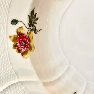 May include: A white porcelain bowl with a scalloped edge and a floral design. The bowl features a yellow and brown poppy with a pink center and green leaves. The bowl has a white woven pattern around the rim.