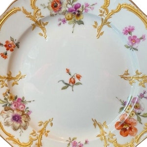 May include: A white porcelain plate with a gold rim and floral pattern. The plate features a variety of flowers in shades of pink, purple, orange, and yellow.