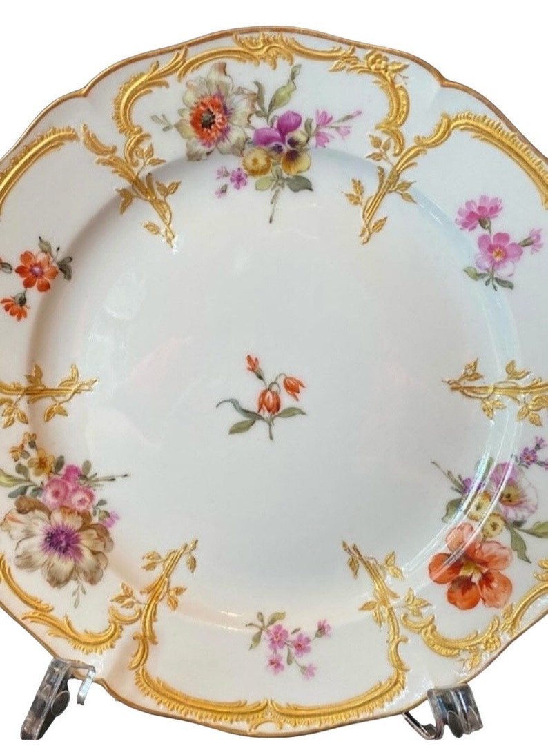May include: A white porcelain plate with a gold rim and floral pattern. The plate features a variety of flowers in shades of pink, purple, orange, and yellow.