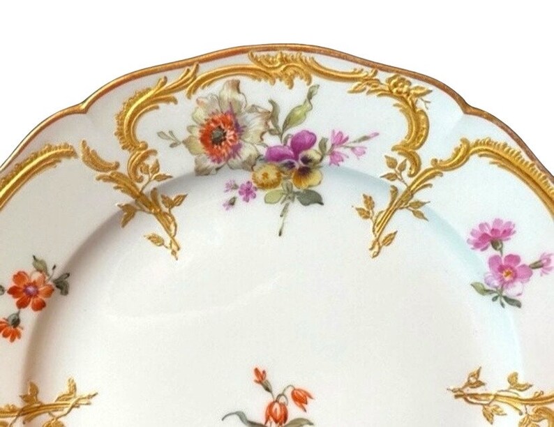 May include: A white porcelain plate with a gold rim and floral pattern. The plate features a delicate floral design with pink, purple, and orange flowers. The gold rim is intricate and adds a touch of elegance to the plate.