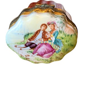 May include: A decorative, antique trinket box with a shell-like shape. The lid features a painted scene of a couple in period clothing, resting on a grassy bank. The box has a gold-colored trim and a reddish-brown top.