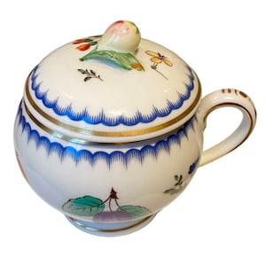 Puede incluir: Un azucarero de porcelana blanca con tapa, con un borde azul y dorado y un diseño floral con frutas. La tapa tiene un pequeño pomo en forma de flor.