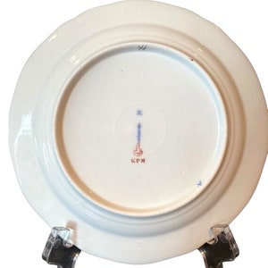 May include: The underside of a white porcelain plate with a blue and red mark that reads "KPM".