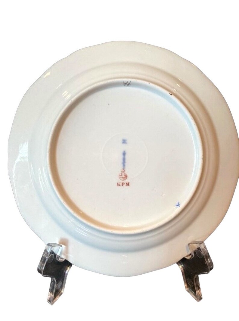 May include: The underside of a white porcelain plate with a blue and red mark that reads "KPM".
