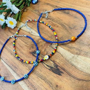 May include: Three beaded necklaces in the image. One is a solid blue, another has a mix of colorful beads, and the third has blue and white floral beads. The necklaces are on a wooden surface.