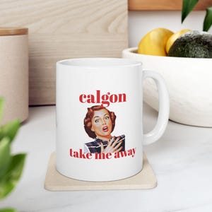 May include: White ceramic mug with a red and white design. The mug features a vintage illustration of a woman with a surprised expression and the text "Calgon take me away".