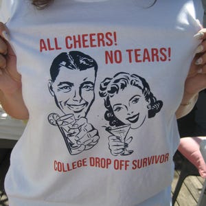 Op de afbeelding: Wit T-shirt met een retro-stijl afbeelding van een man en een vrouw die drankjes vasthouden. De tekst luidt "All Cheers! No Tears! College Drop Off Survivor".