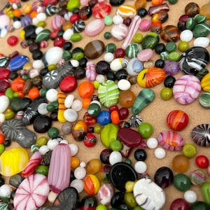 May include: A collection of colourful glass beads in various shapes and sizes. The beads are arranged on a brown surface and include swirls, stripes, and solid colours.