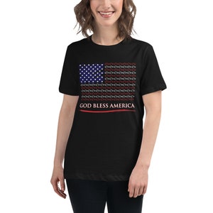 Op de afbeelding: Zwart T-shirt met een Amerikaans vlagontwerp bestaande uit witte sterren en rode, witte en blauwe vissymbolen. De tekst "GOD BLESS AMERICA" staat eronder in het wit gedrukt, met een rode lijn eronder.