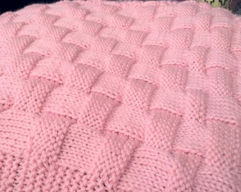 May include: A pink knitted blanket with a woven pattern. The blanket is made of soft yarn and has a delicate texture.