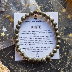 May include: A pyrite gemstone bracelet with metallic gray beads. The bracelet is displayed on a card with the text "Rainy Day Apothecary PYRITE" and information about the stone's properties. The background includes a celestial map and crystals.